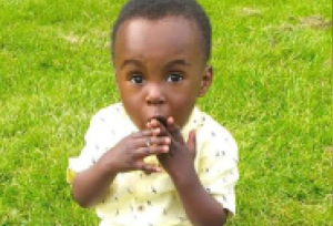 Photograph of Awaab Ishak sitting on grass in a little shirt and trousers, provided to the press by his family.